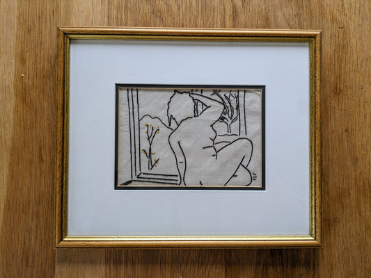 Handstitched image of a nude woman seated looking out of a window. The image is shown in  a wooden and gold painted frame with a cream mount.